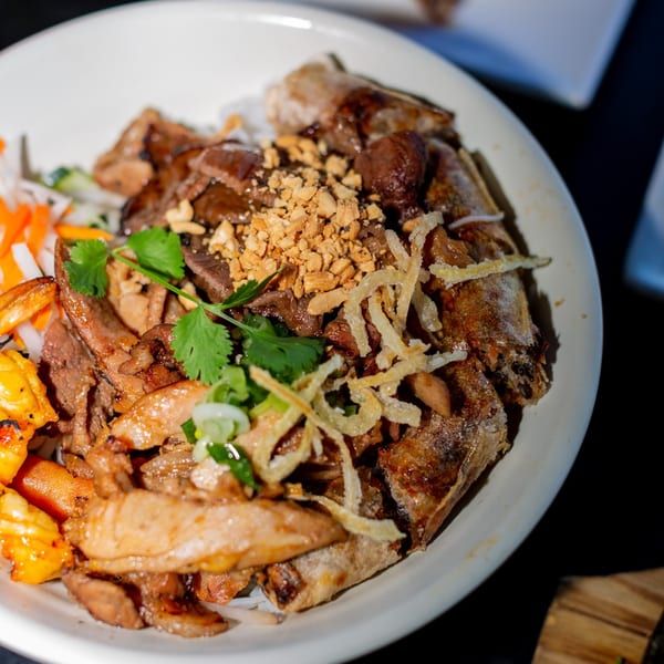 A Vietnamese vermicelli bowl with grilled meats, fried spring rolls, and vegetables, topped with peanuts.