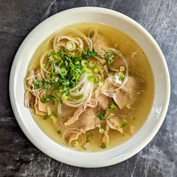 Now Phở's beef pho with broth, sliced onions, and fresh herbs in a white bowl.